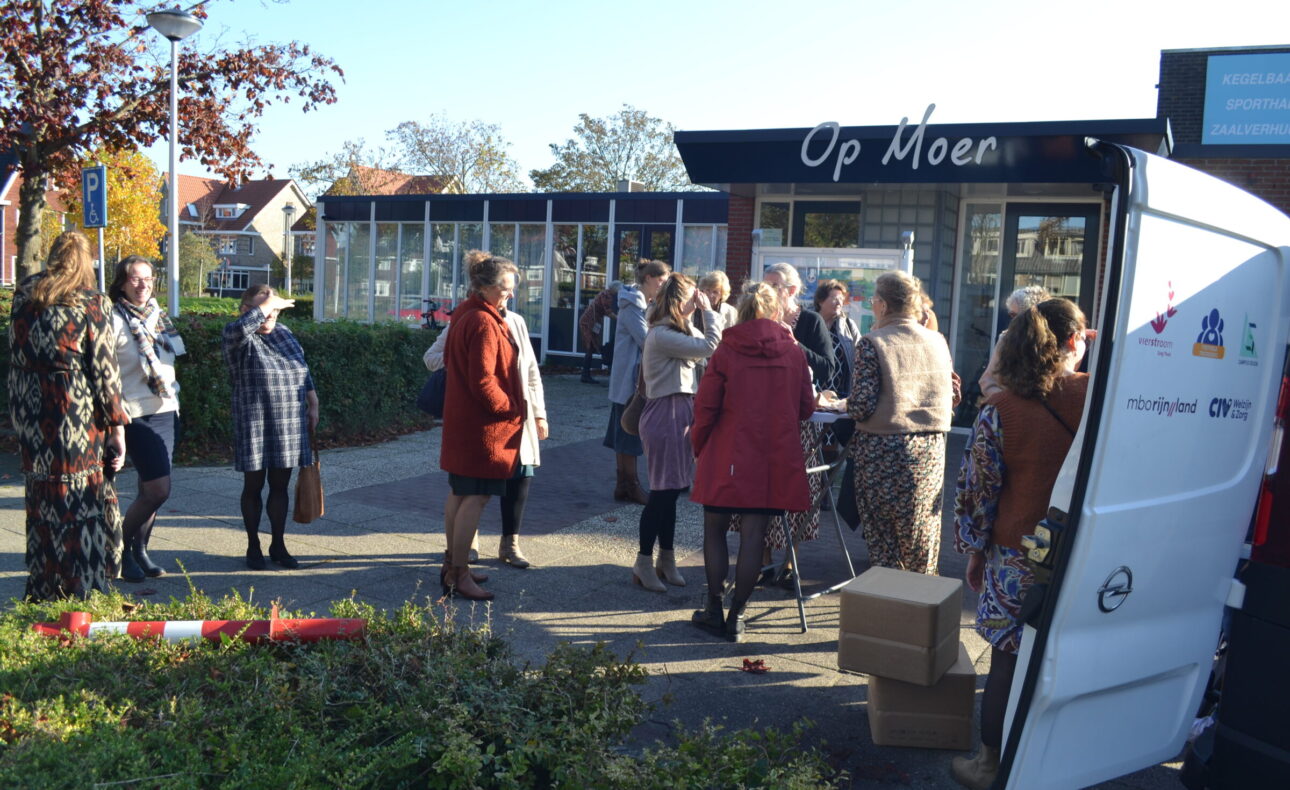 Groep mensen verzameld bij een busje voor een gebouw met de naam "Op Moer". Het is een zonnige dag met blauwe lucht.
