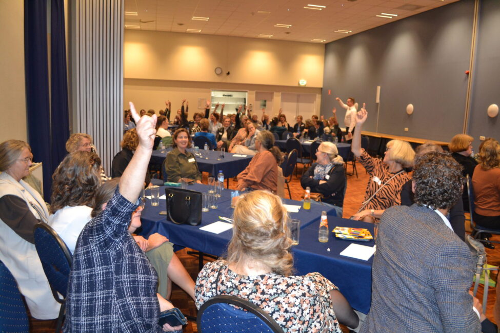Groep mensen zit aan tafels in een zaal, sommigen steken hun hand op. De ruimte heeft blauwe muren en een houten vloer.