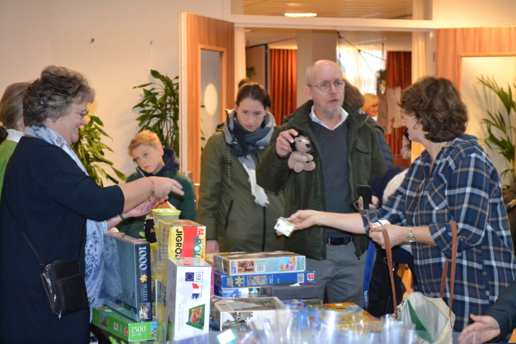 Groep mensen verzameld rond een tafel met bordspellen in een binnenruimte. Een vrouw reikt een object aan een man aan.