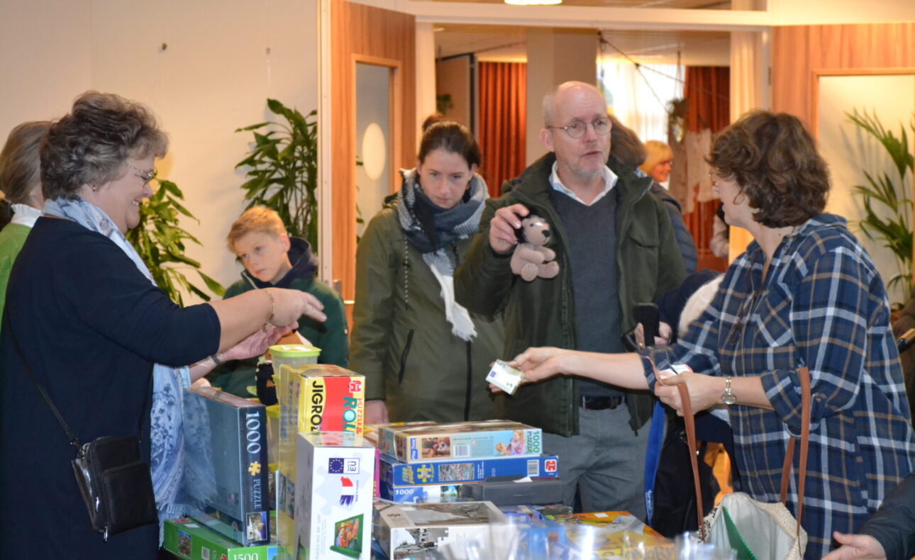 Groep mensen verzameld rond een tafel met bordspellen in een binnenruimte. Een vrouw reikt een object aan een man aan.