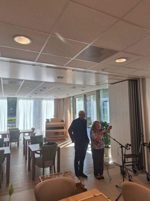 Twee mensen staan in een lichte kamer met tafels en stoelen, terwijl ze samen naar iets kijken. Er is veel natuurlijk licht.