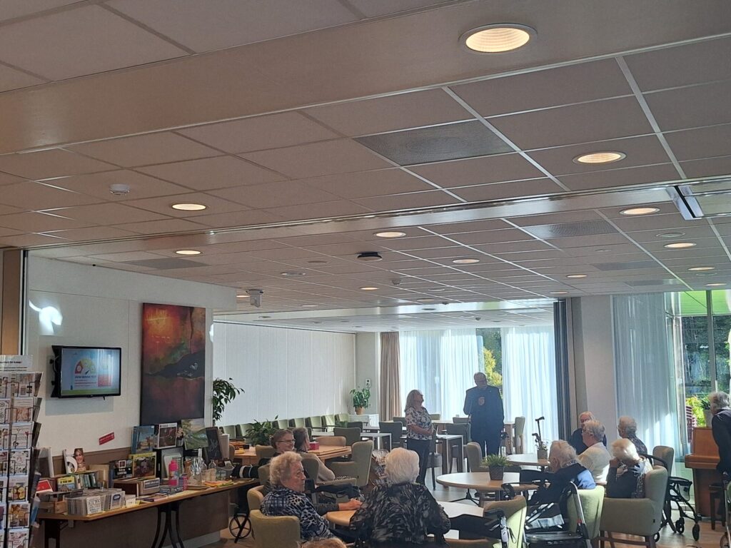Een groep ouderen zit in een gemeenschappelijke ruimte met tafels en stoelen, terwijl ze naar een spreker luisteren.