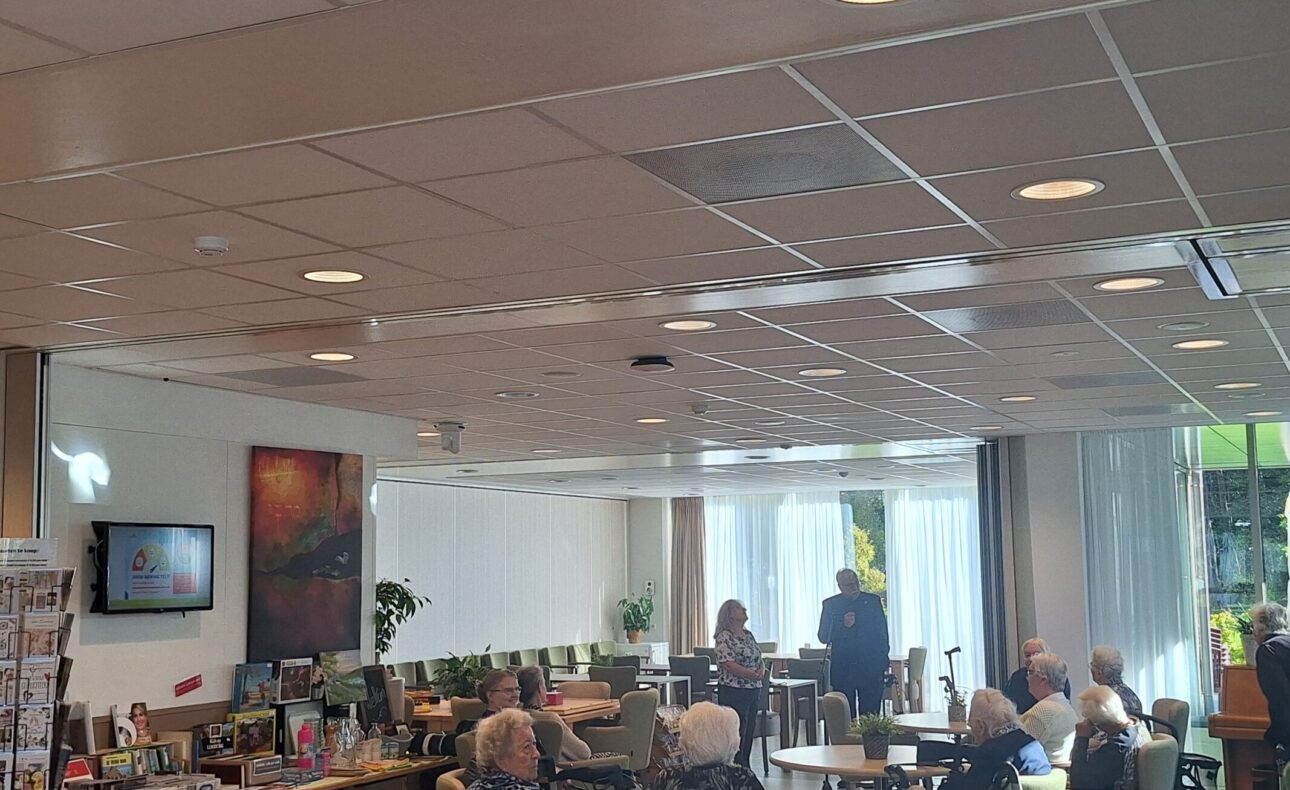 Een groep ouderen zit in een gemeenschappelijke ruimte met tafels en stoelen, terwijl ze naar een spreker luisteren.
