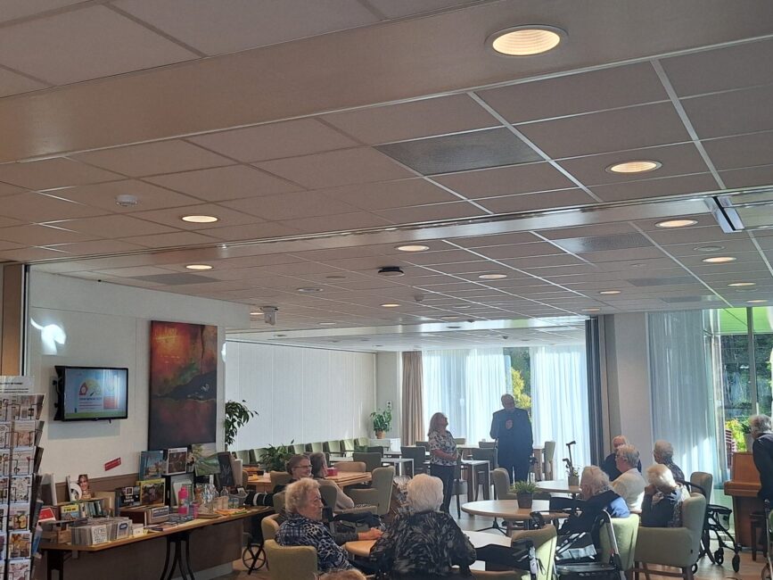 Een groep ouderen zit in een gemeenschappelijke ruimte met tafels en stoelen, terwijl ze naar een spreker luisteren.