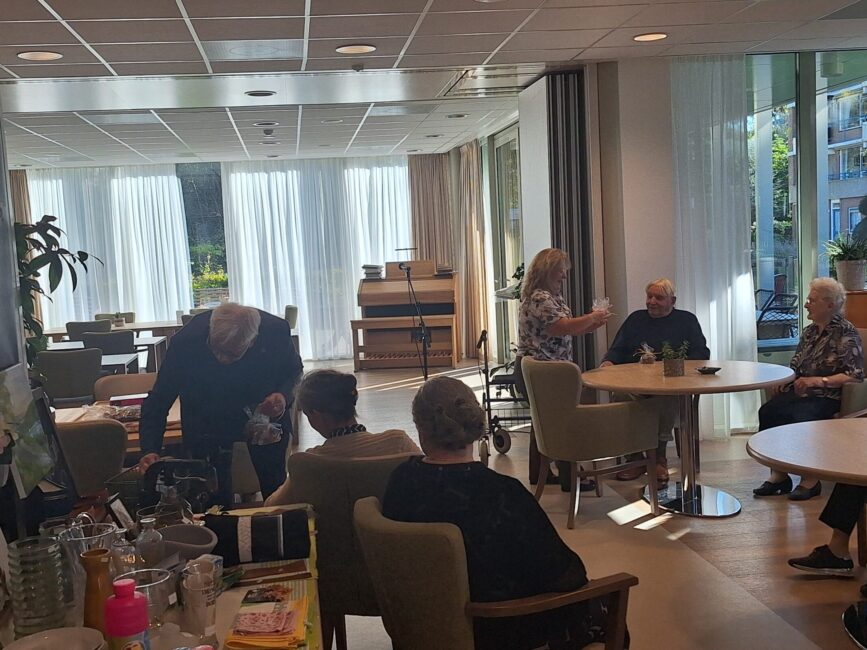 Een groep ouderen zit samen in een lichte ruimte met grote ramen. Er is een tafel met drankjes en een vrouw die serveert.