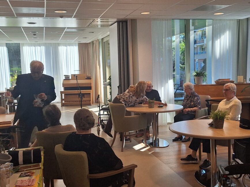 Groep ouderen zit samen in een lichte ruimte met tafels en stoelen, terwijl een man voor hen staat en iets presenteert.
