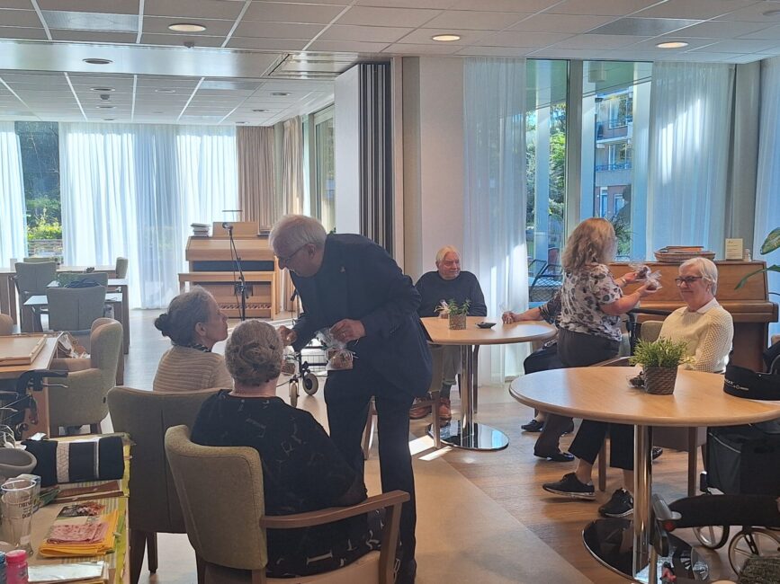 Een groep ouderen geniet van een gezellige bijeenkomst in een lichte ruimte met grote ramen en tafels, op 5 oktober 2024.