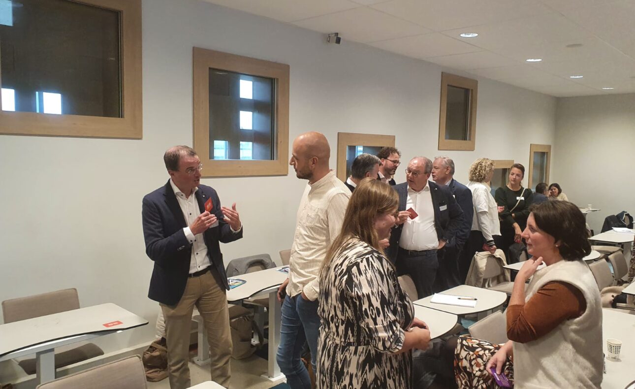 Een groep mensen staat in een vergaderruimte te praten. Ze dragen formele kleding en lijken in een levendige discussie verwikkeld.
