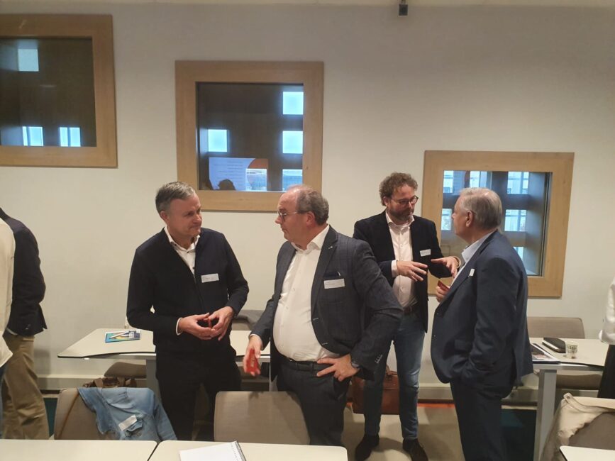 Vier mannen in gesprek in een vergaderruimte met tafels en stoelen. Twee mannen dragen een jasje, de anderen een trui.