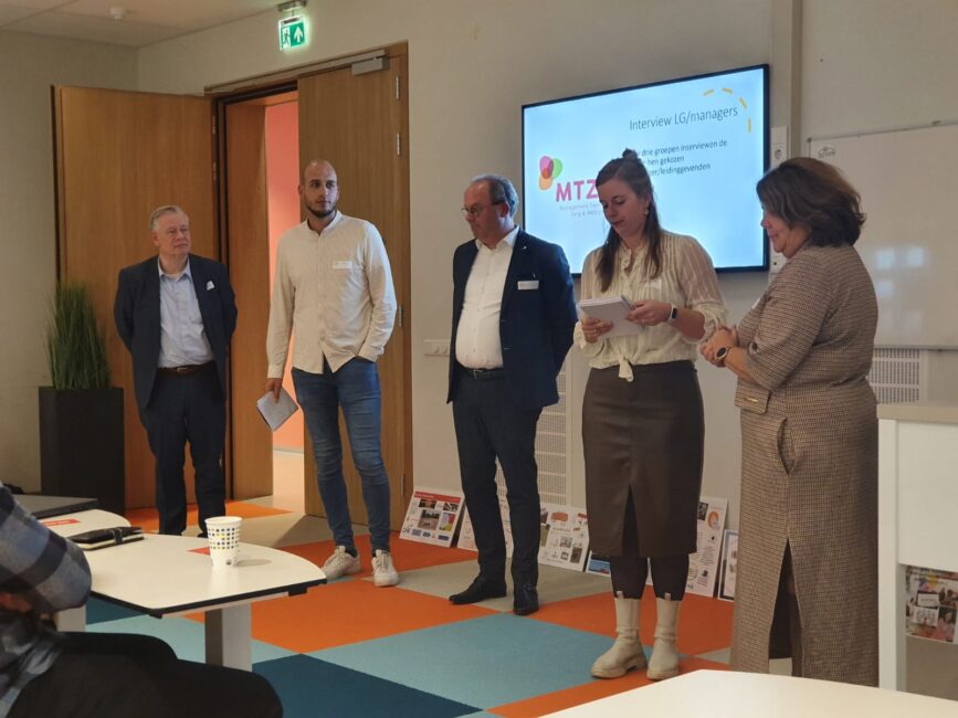 Vijf mensen staan in een vergaderruimte voor een scherm, in gesprek. Er liggen tijdschriften op de tafel en vloer.