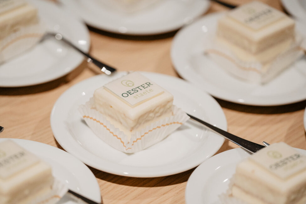 Borden met vierkante gebakjes, elk met een vorkje. Op de gebakjes staat "Oester" gedrukt. Ze staan op een houten tafel.