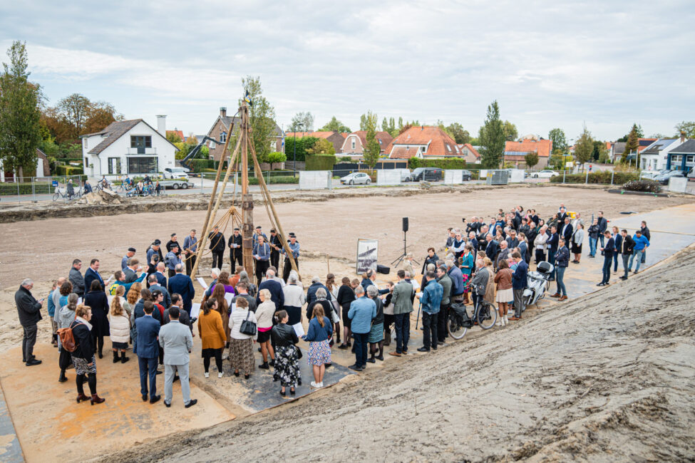 Groep mensen verzameld op een bouwplaats tijdens een ceremonie. Op de achtergrond zijn huizen en bomen zichtbaar.