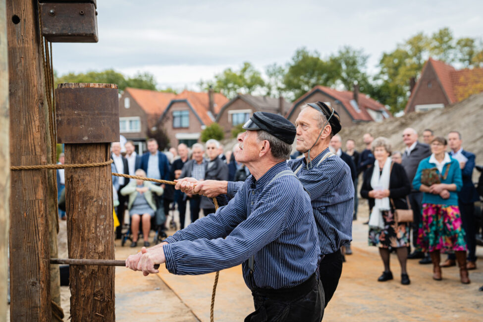 Twee mannen in traditionele kleding trekken aan touwen tijdens een evenement, met een groep toeschouwers op de achtergrond.