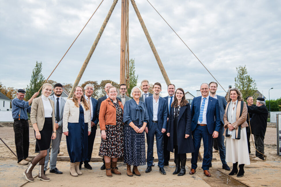 Groep mensen poseert voor een houten constructie op een bouwplaats. Ze dragen nette kleding en lachen naar de camera.