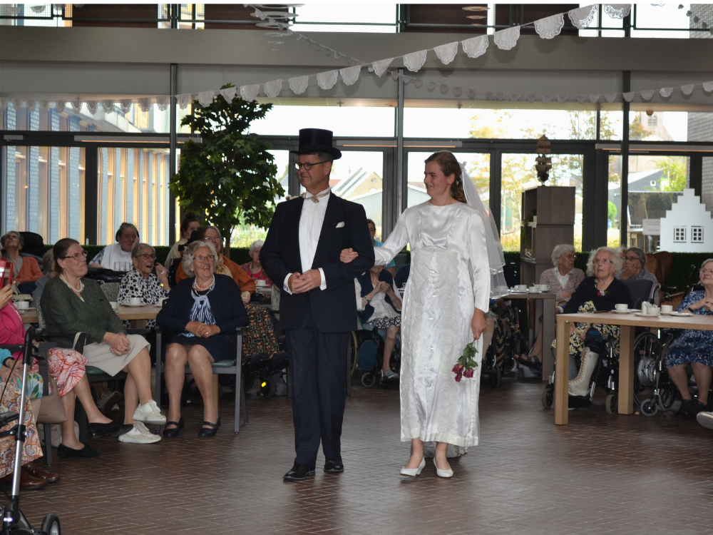 Een bruidspaar loopt over de catwalk tijdens een bruidsmodeshow, omringd door toeschouwers in een zaal met grote ramen.