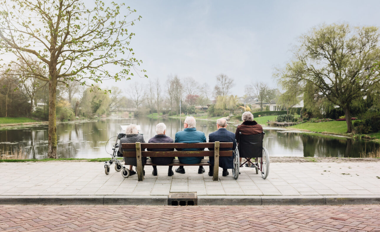 Vijf ouderen zitten op een bankje aan het water, omringd door bomen en een rustige omgeving op een lentedag.