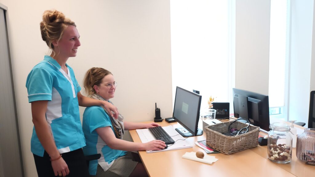Twee vrouwen in blauwe uniformen werken samen aan een bureau met computers in een kantooromgeving. Er staat een mand op tafel.