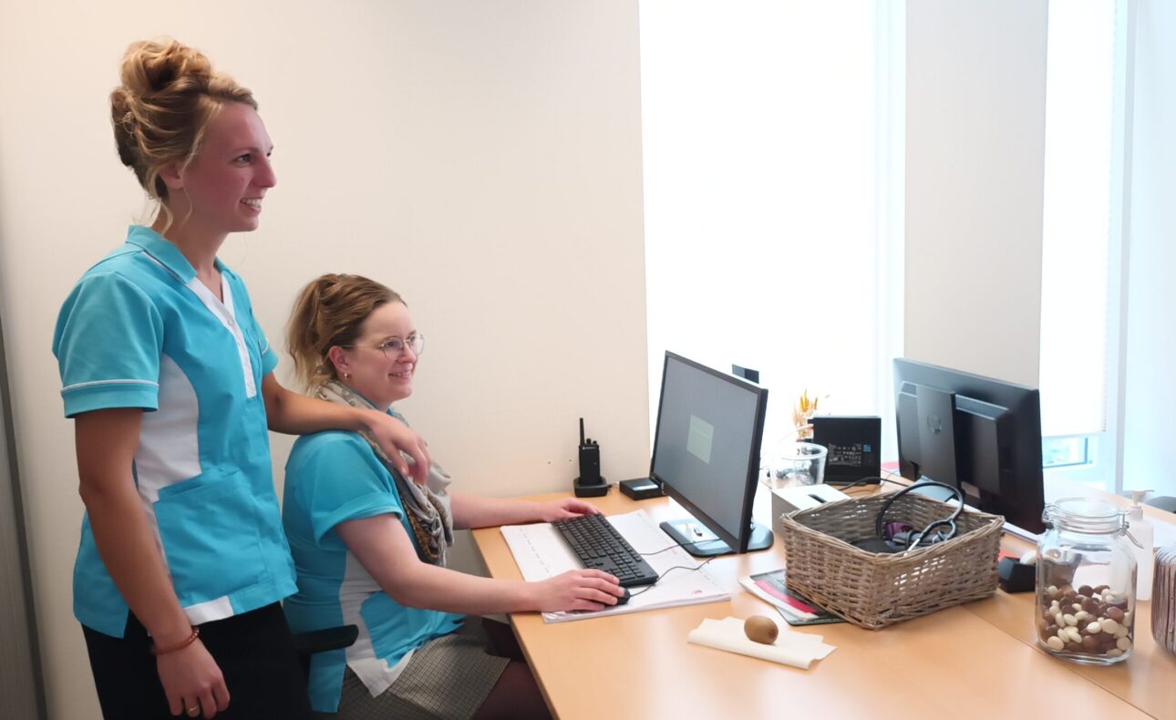 Twee vrouwen in blauwe uniformen werken samen aan een bureau met computers in een kantooromgeving. Er staat een mand op tafel.