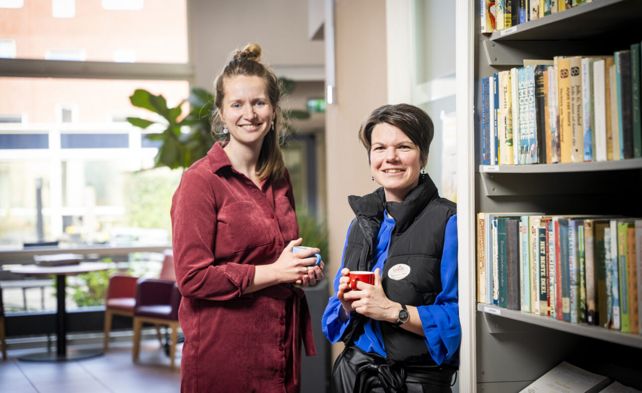 Twee vrouwen staan glimlachend in een bibliotheek, elk met een mok in de hand. Op de achtergrond zijn boekenplanken zichtbaar.