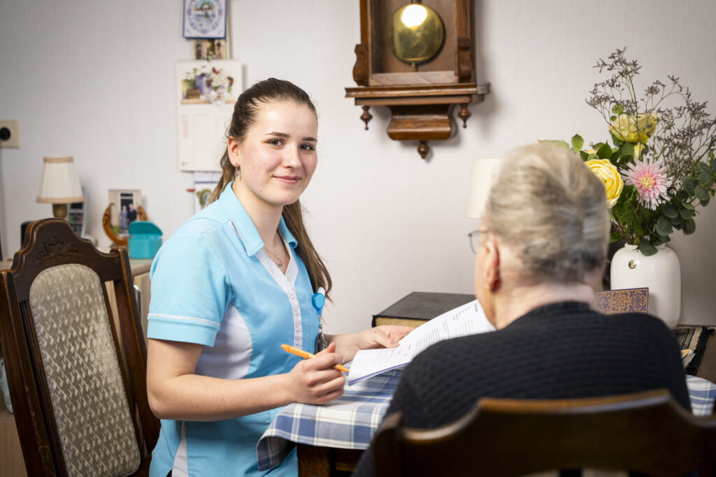 Een zorgverlener in een blauw uniform zit aan tafel met een oudere vrouw. Ze glimlacht en houdt een pen vast bij een notitieblok.