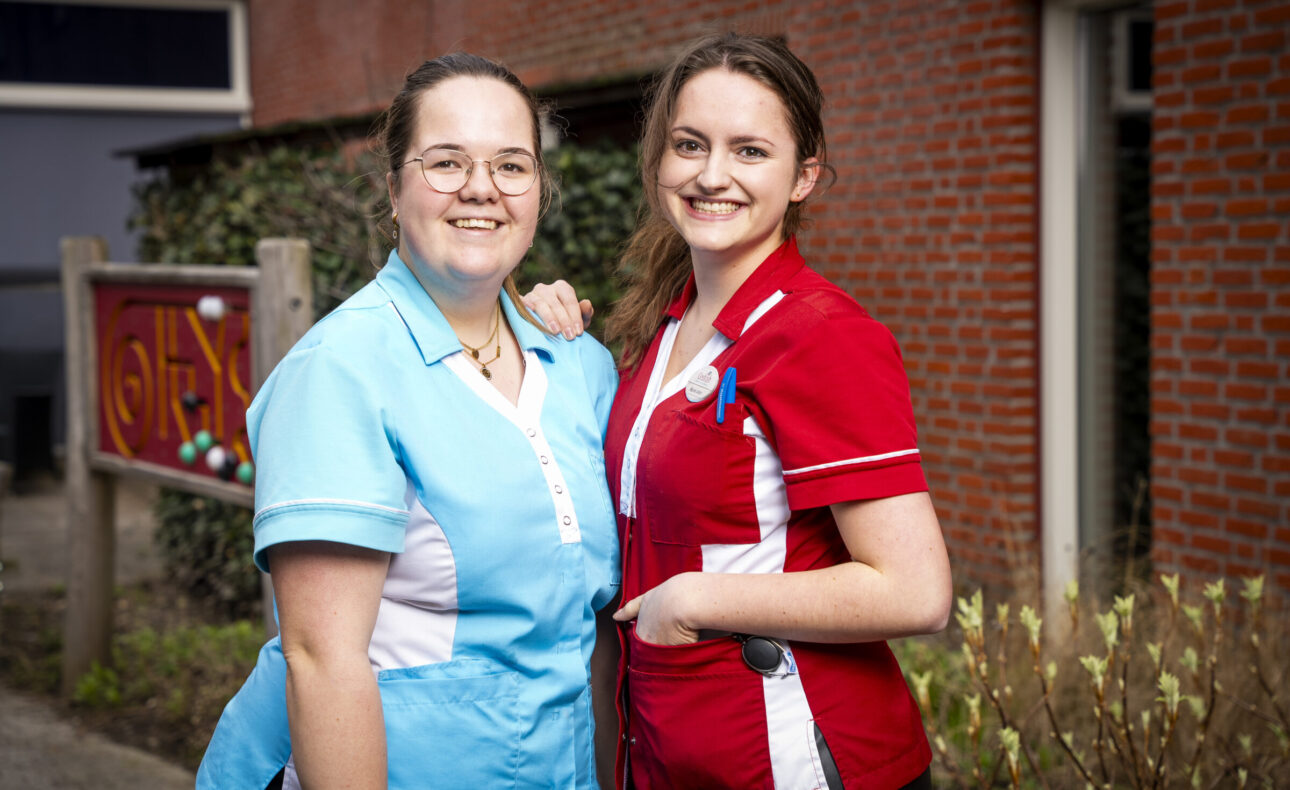 Twee zorgmedewerkers in blauwe en rode uniformen staan glimlachend buiten voor een bakstenen gebouw met een bord op de achtergrond.