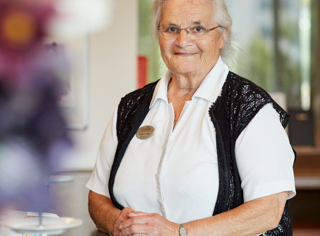 Oudere vrouw met bril en wit overhemd, staand bij een balie in een lichte ruimte. Op de voorgrond zijn bloemen en fruit.