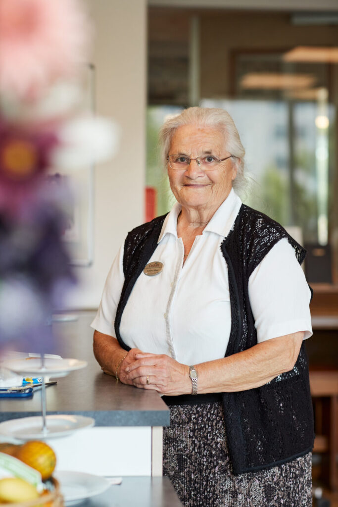 Oudere vrouw met bril en wit overhemd, staand bij een balie in een lichte ruimte. Op de voorgrond zijn bloemen en fruit.