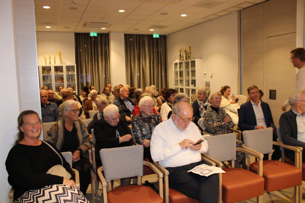 Groep oudere mensen zit in een zaal met stoelen, sommigen praten en anderen kijken naar hun telefoon of een boekje.