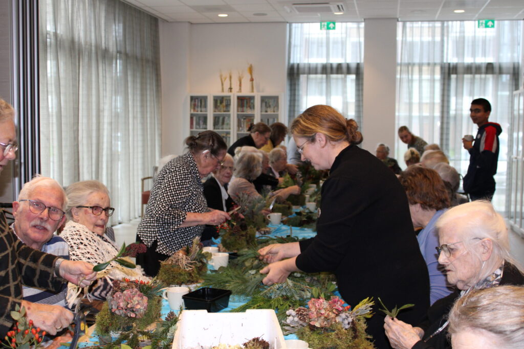 Groep ouderen en begeleiders maken bloemstukken aan een lange tafel in een lichte ruimte met grote ramen.