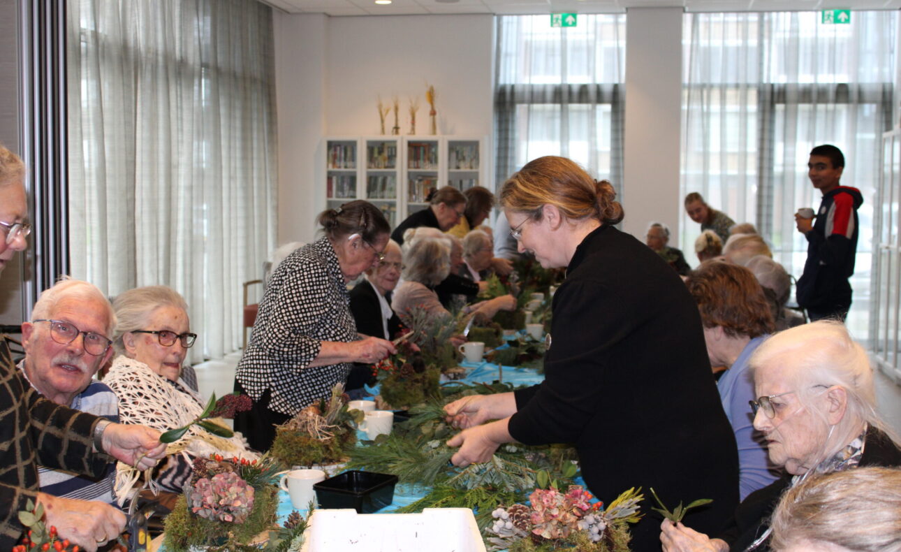 Groep ouderen en begeleiders maken bloemstukken aan een lange tafel in een lichte ruimte met grote ramen.