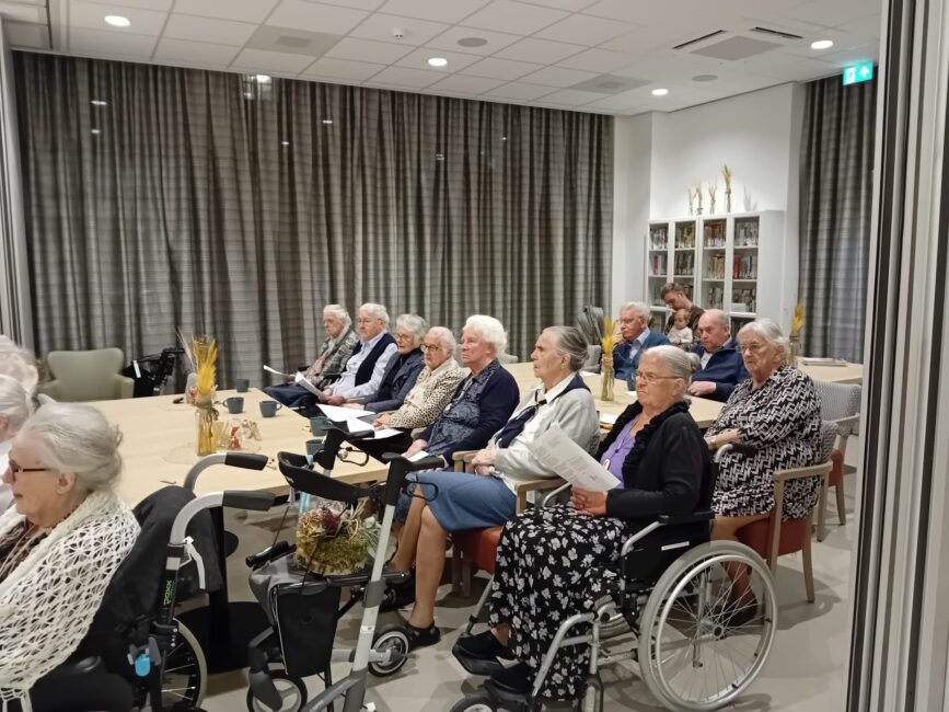 Een groep ouderen zit in een zaal, aandachtig luisterend naar een optreden. Sommigen zitten in rolstoelen, anderen op stoelen.