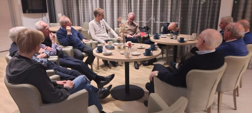 Een groep mannen zit in een kring in een gezellige ruimte, pratend en genietend van koffie en thee op een bijeenkomst.
