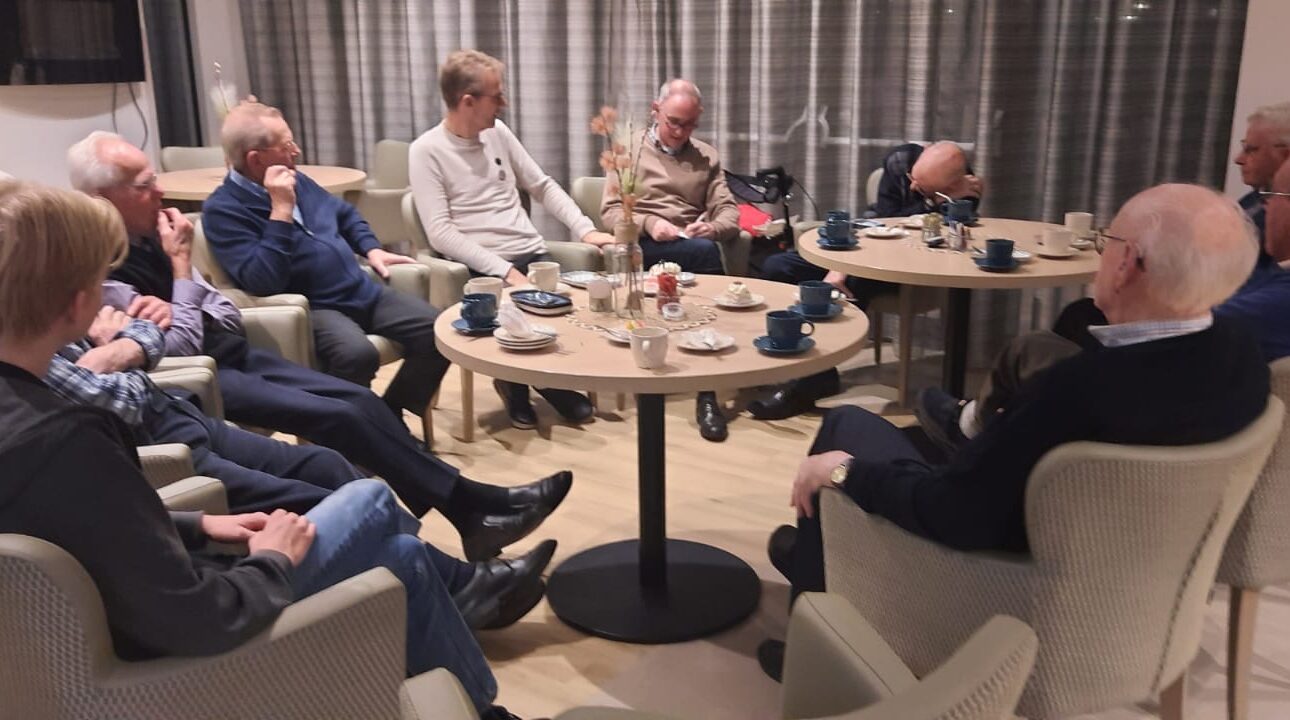 Een groep mannen zit in een kring in een gezellige ruimte, pratend en genietend van koffie en thee op een bijeenkomst.