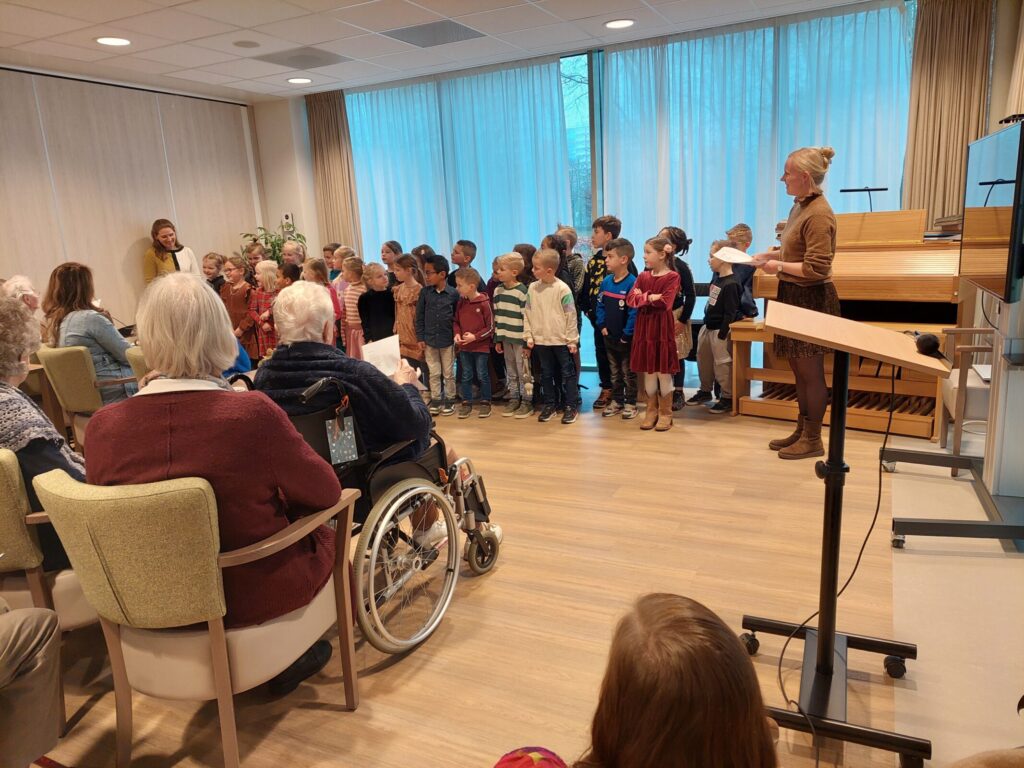 Een groep kinderen zingt voor een publiek van ouderen in een zaal. Een volwassene begeleidt hen met een muziekstandaard.