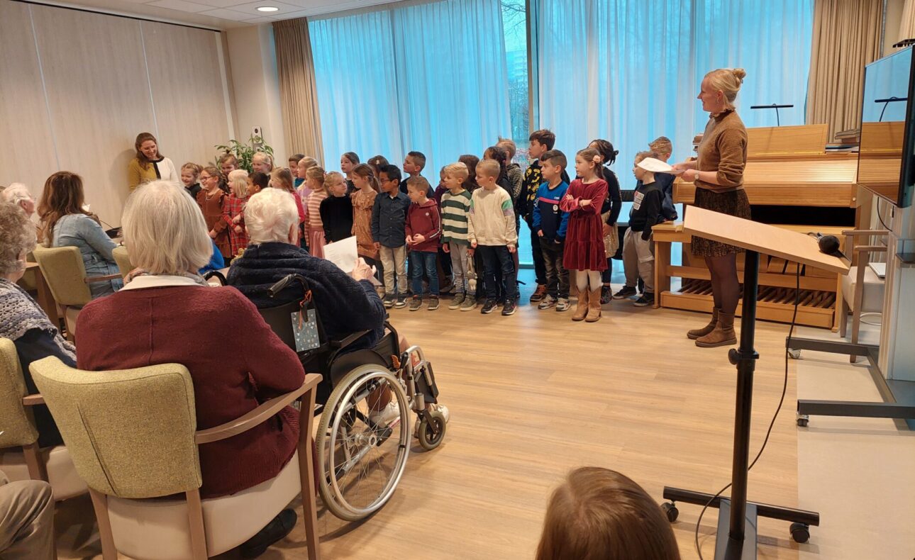 Een groep kinderen zingt voor een publiek van ouderen in een zaal. Een volwassene begeleidt hen met een muziekstandaard.