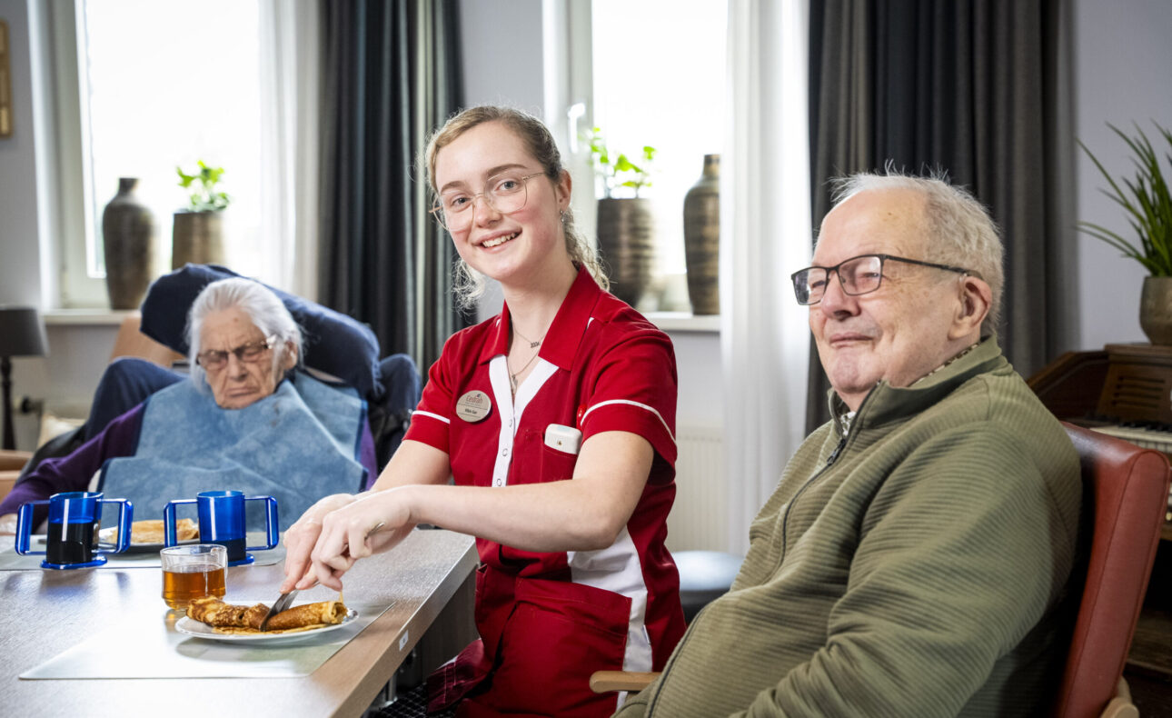 Een zorgmedewerker in rood uniform zit glimlachend aan tafel met twee oudere mensen in een gezellige kameromgeving.