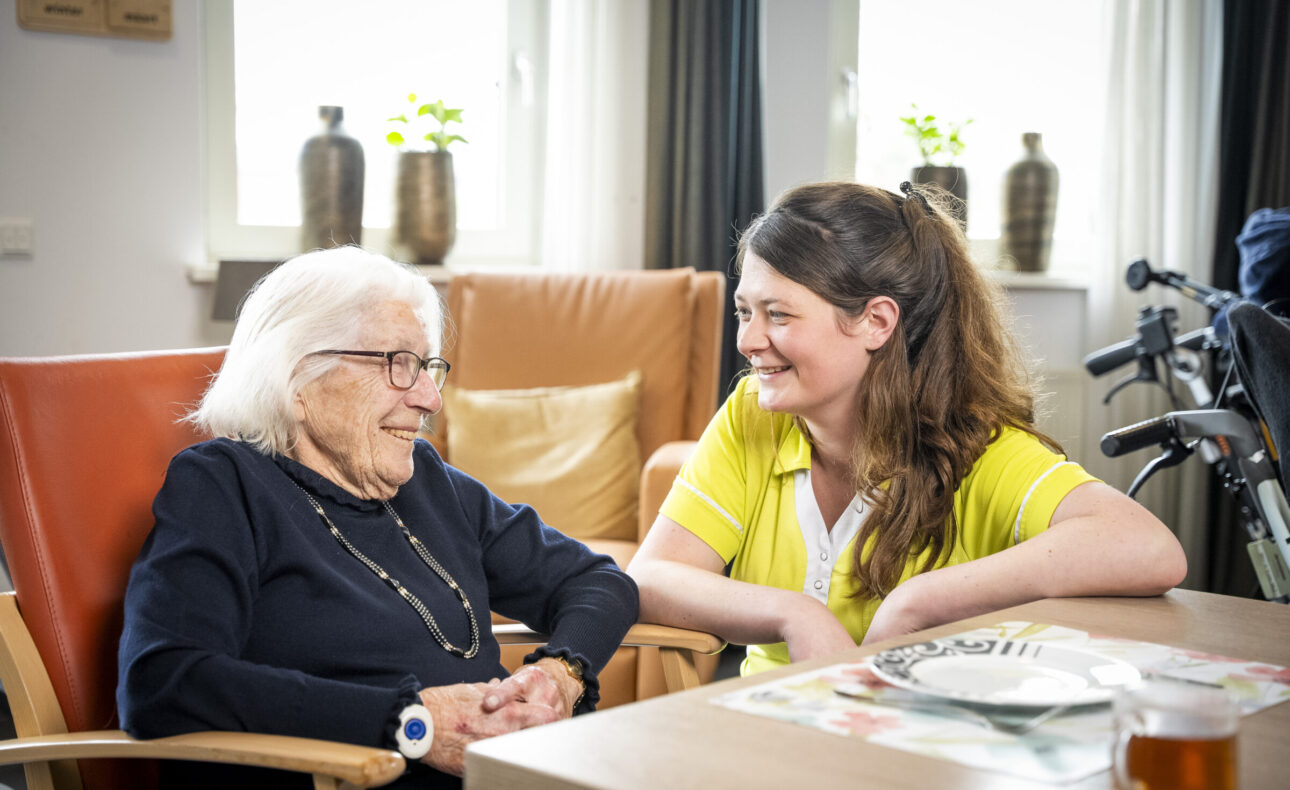 Twee vrouwen zitten glimlachend aan een tafel in een zonnige kamer. Een oudere vrouw en een jongere vrouw in een geel uniform.