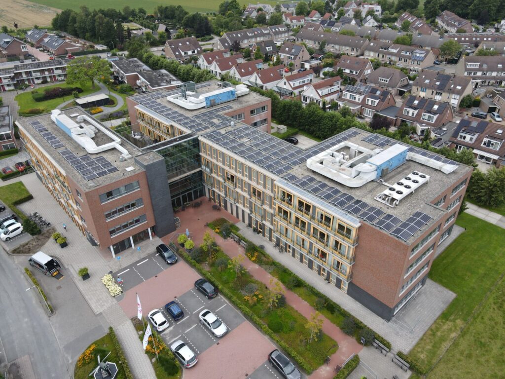 Luchtfoto van een groot gebouw met zonnepanelen op het dak, omringd door een woonwijk en groene gebieden in de omgeving.