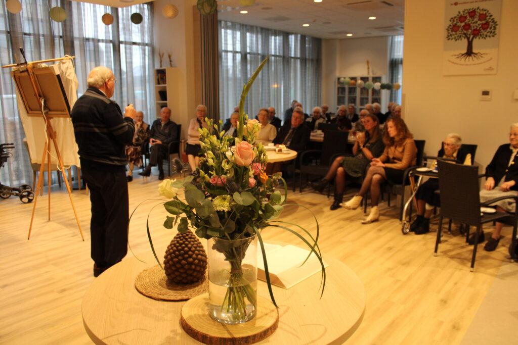 Een oudere man geeft een presentatie aan een groep mensen in een zaal. Op de voorgrond staat een vaas met bloemen.