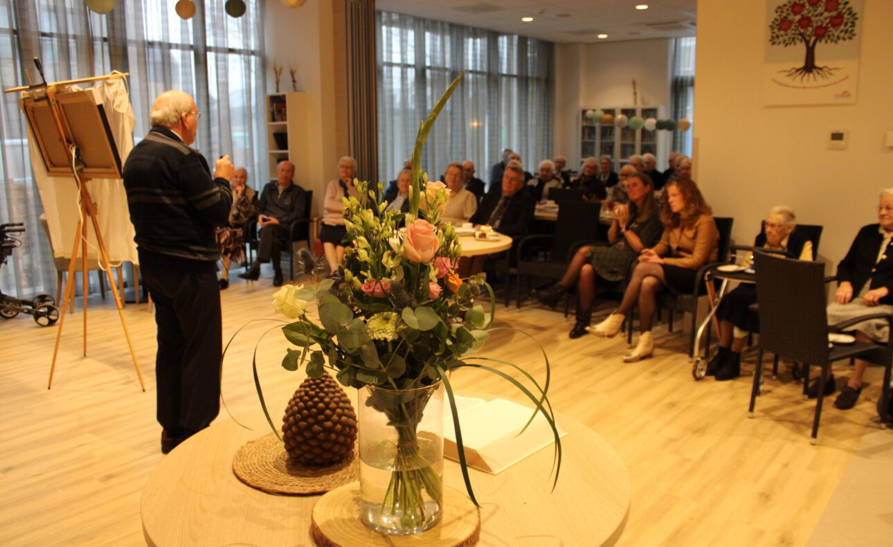 Een oudere man geeft een presentatie aan een groep mensen in een zaal. Op de voorgrond staat een vaas met bloemen.