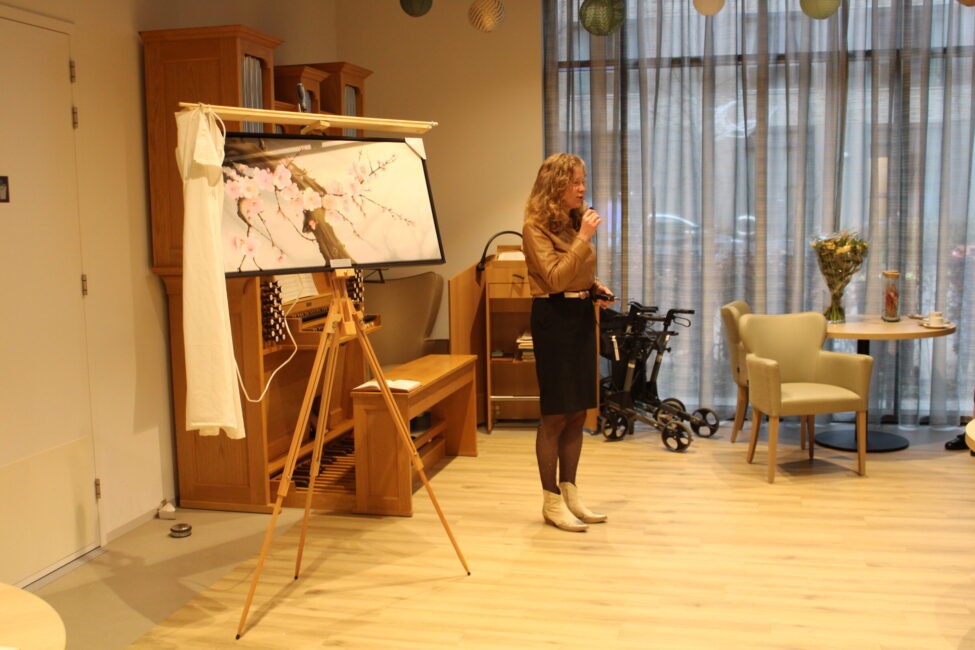 Vrouw geeft presentatie in een kamer met houten vloer, schildersezel met kunstwerk, en meubels. Grote ramen op de achtergrond.