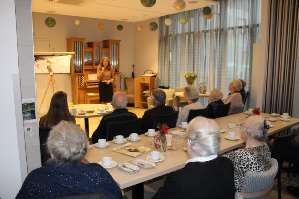 Een groep ouderen zit aan een tafel met koffie en gebak, terwijl een vrouw vooraan een presentatie geeft in een lichte ruimte.