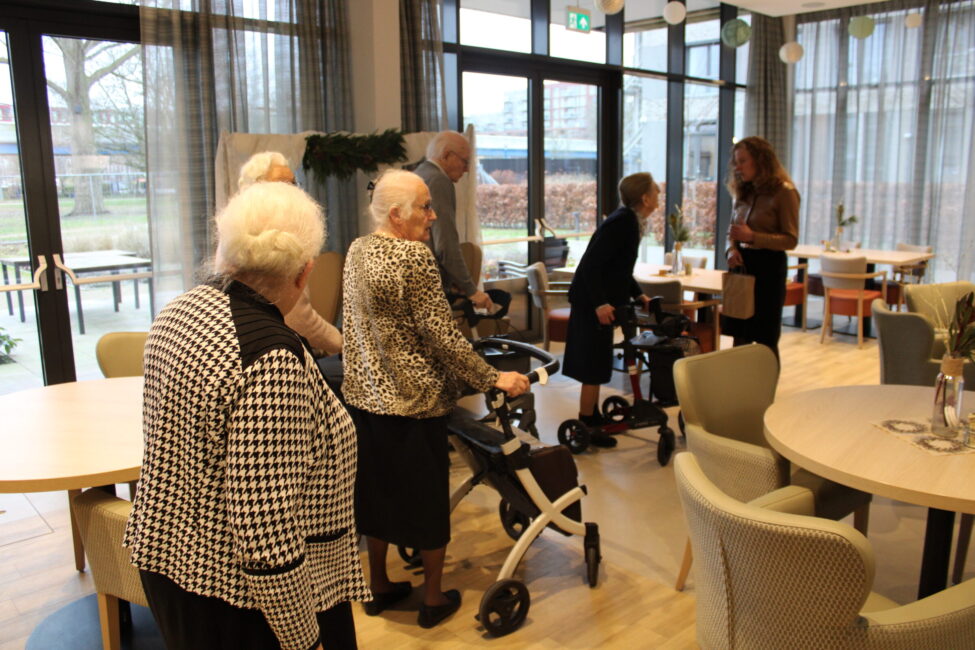 Groep ouderen in een gemeenschappelijke ruimte, sommigen met rollators. Ze praten met een vrouw bij een tafel in een verzorgingshuis.