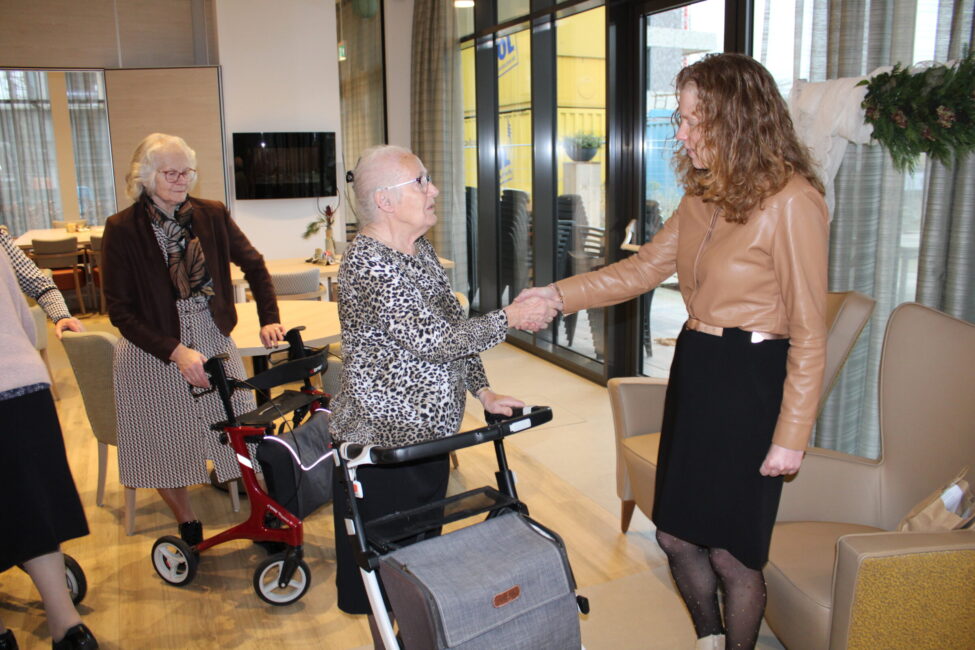 Twee vrouwen schudden elkaar de hand in een verzorgingstehuis, terwijl een andere vrouw met een rollator toekijkt.