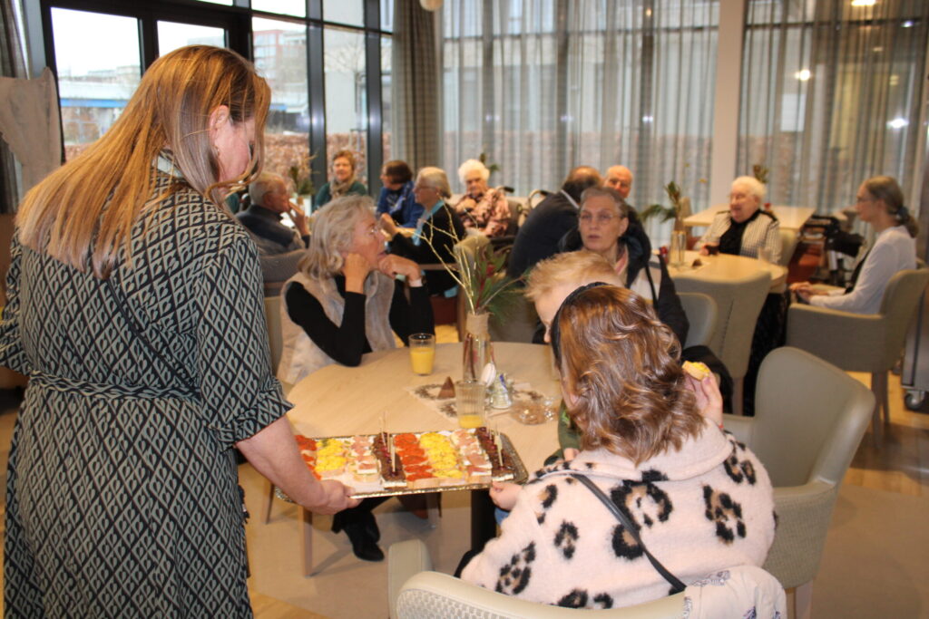 Een vrouw serveert een schaal met gebak aan een groep mensen die aan tafels zitten in een gezellige, lichte ruimte.