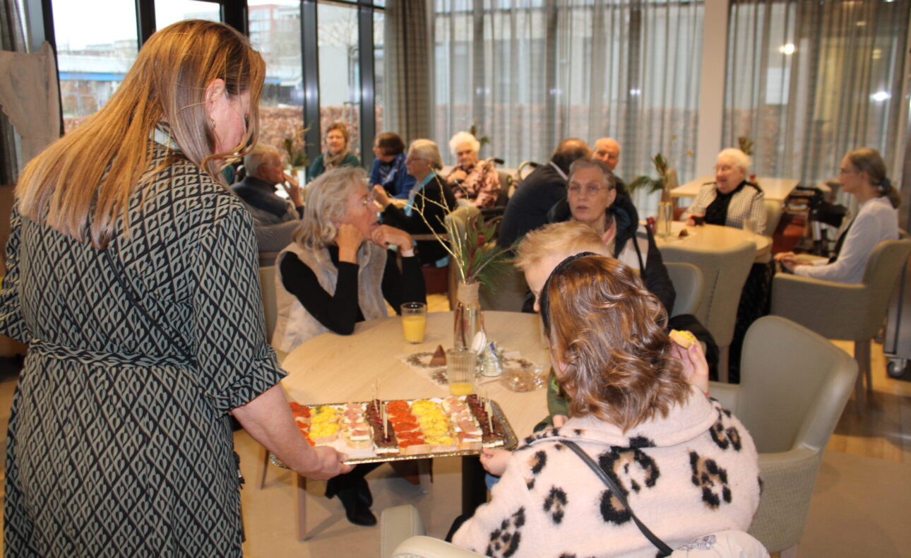 Een vrouw serveert een schaal met gebak aan een groep mensen die aan tafels zitten in een gezellige, lichte ruimte.