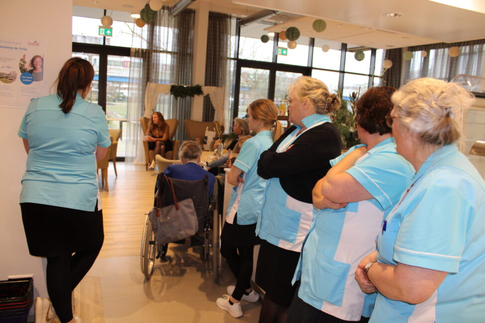 Groep zorgmedewerkers in blauwe uniformen kijkt naar een presentatie in een gemeenschappelijke ruimte van een zorginstelling.