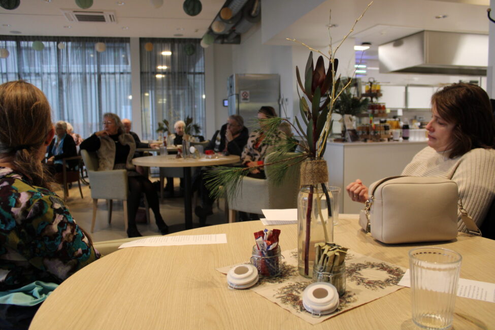 Een groep mensen zit aan tafels in een gezellige ruimte met planten en decoraties. Op de voorgrond staat een vaas met bloemen.