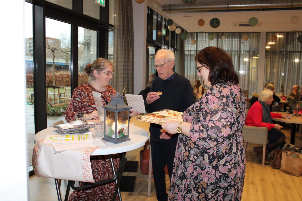 Drie mensen staan bij een tafel met een lantaarn en hapjes in een gezellige ruimte met grote ramen en gordijnen.