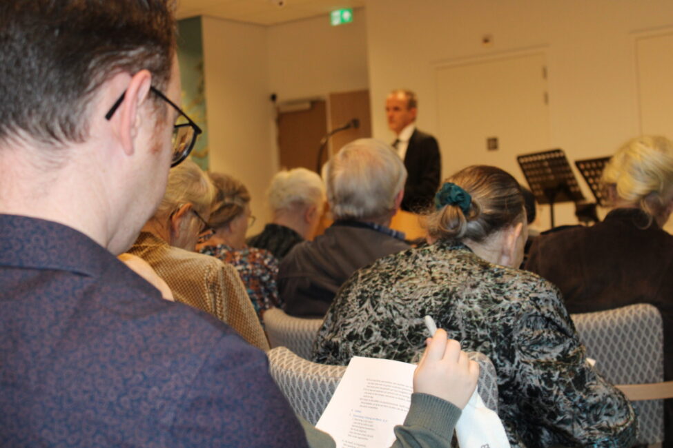 Een groep mensen zit in een zaal, aandachtig luisterend naar een spreker die vooraan staat. Een persoon maakt aantekeningen.